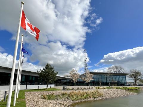 Flags flying outside SKAO Global HQ. The focus is on the Canadian flag, with the building and a small lake behind.