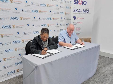 A picture of Prof. Phil Diamond, Director-General of the SKAO and Pontscho Maruping, managing director of SARAO, each signing a document while sitting at a table. The wall behind them shows logo’s of AfAS, SARAO and the IAU, To the right of the table stands a banner with the SKAO logo and some numbers on the SKA-Mid telescope.