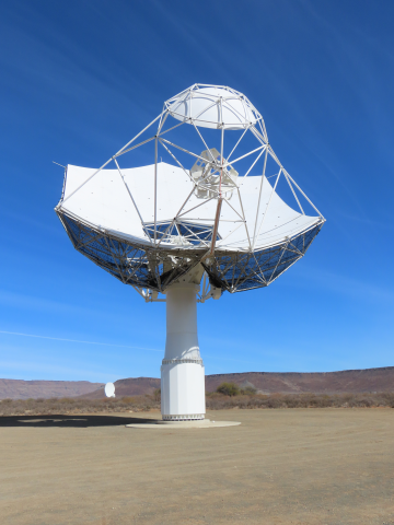 Picture of the first SKA-Mid dish against a blue sky. The dish has a hexagonal shape and sits atop a white pedestal. A smaller spherical shaped mirror is supported by small white bars.  A triangular structure, the feed indexer, is also mounted on these bars.