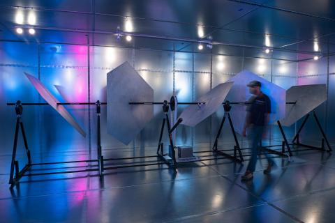 A picture of the SKA-Low reverberation chamber. The rectangular room is made from iron panelling. Inside sit different hexagonal-shaped panels in various angles attached to a black frame. A man walks next to the hexagonal shapes, which shows that the height of the ceiling is as high as any normal room.  