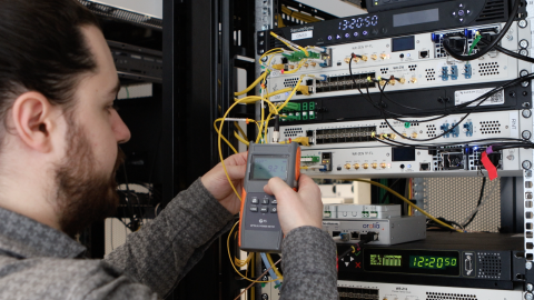 A picture of a man holding an electrical meter connected with wires to electrical equipment in the background. 