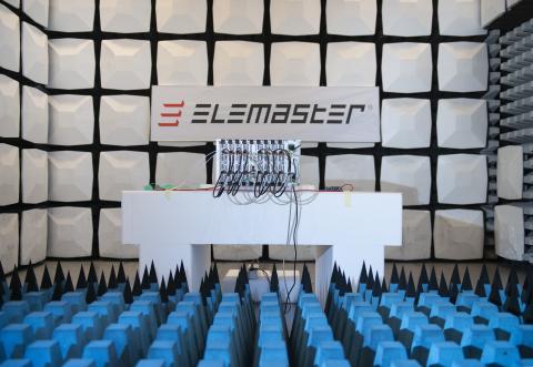 A picture of an electronic unit on top of a white bench in a testing room. A logo of Elemaster hangs against the back panelling above the unit. The floor is covered in blue foam pieces in the form of cut-off pyramids. 