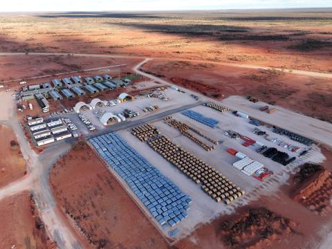 Aerial picture of the SKA-Low construction camp. The camp consists of several tents and has hundreds of components of construction material in neat stacks laid out on top of a grey underground. This colour contrasts with the nature surrounding the camp. 