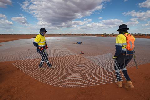 Two men in high vis clothing carrying a sheet of iron mesh ready to place it down to tens of other sheets. The mesh lies on top of the ground with a blue sky overhead.  