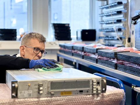 A man wearing gloves dusting of an SKA-Mid digitiser. The digitiser looks like a grey desktop computer lying on its side. In the background several other digitisers are lying on a table wrapped in protective insulation.