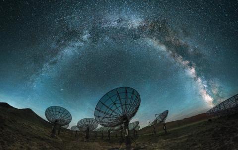 Telescopes dishes during the night time. The Milky Way crosses the sky as a bright arc with darker cloud-like structures.