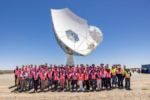 Group picture of people in mostly pink high vis vests in front of a telescope dish with clear blue skies overhead.