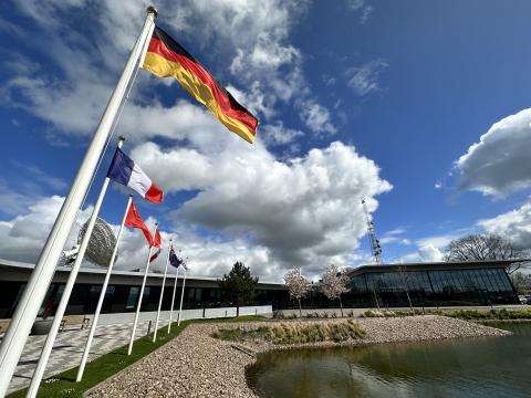 Picture of country flags waving next to a pond and a building. The front flag is that of Germany followed by the French and Chinese flag. The blue sky is filled with big fluffy clouds.