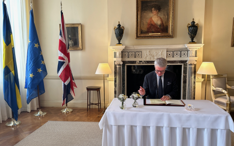 Swedish Ambassador Stefan Gullgren signing the SKAO Convention. He is sitting at a table in front of a fireplace. In the background are the Swedish, EU and UK flags.